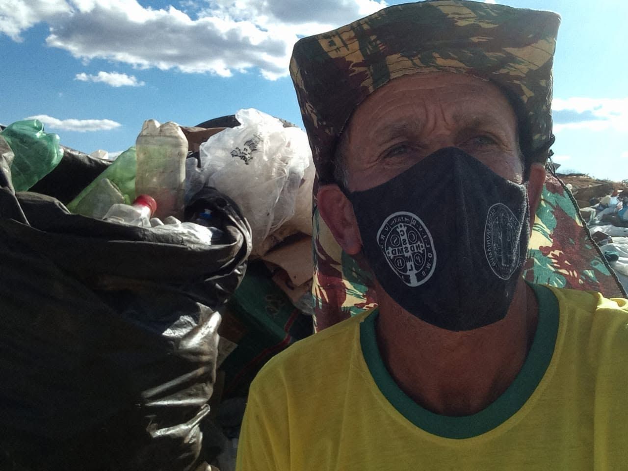 Catador de máscara e chapéu ao lado de sacos de recicláveis em lixão